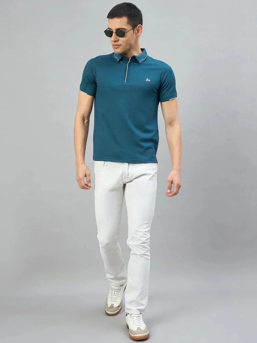 Man in 98degreenorth Men's Teal Polo T-Shirt with LA Logo, white jeans, and sneakers on a plain grey background.