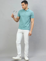 Man in a 98degreenorth turquoise quarter zip polo t-shirt and white jeans, standing against a gray background.
