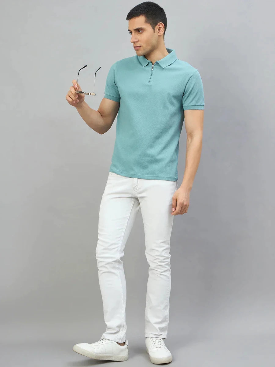 Man in a 98degreenorth turquoise quarter zip polo t-shirt and white jeans, standing against a gray background.
