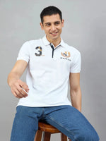 A man in a 98degreenorth Men's White AR Elite Club No.3 Polo T-Shirt sits on a stool, smiling against a gray background.