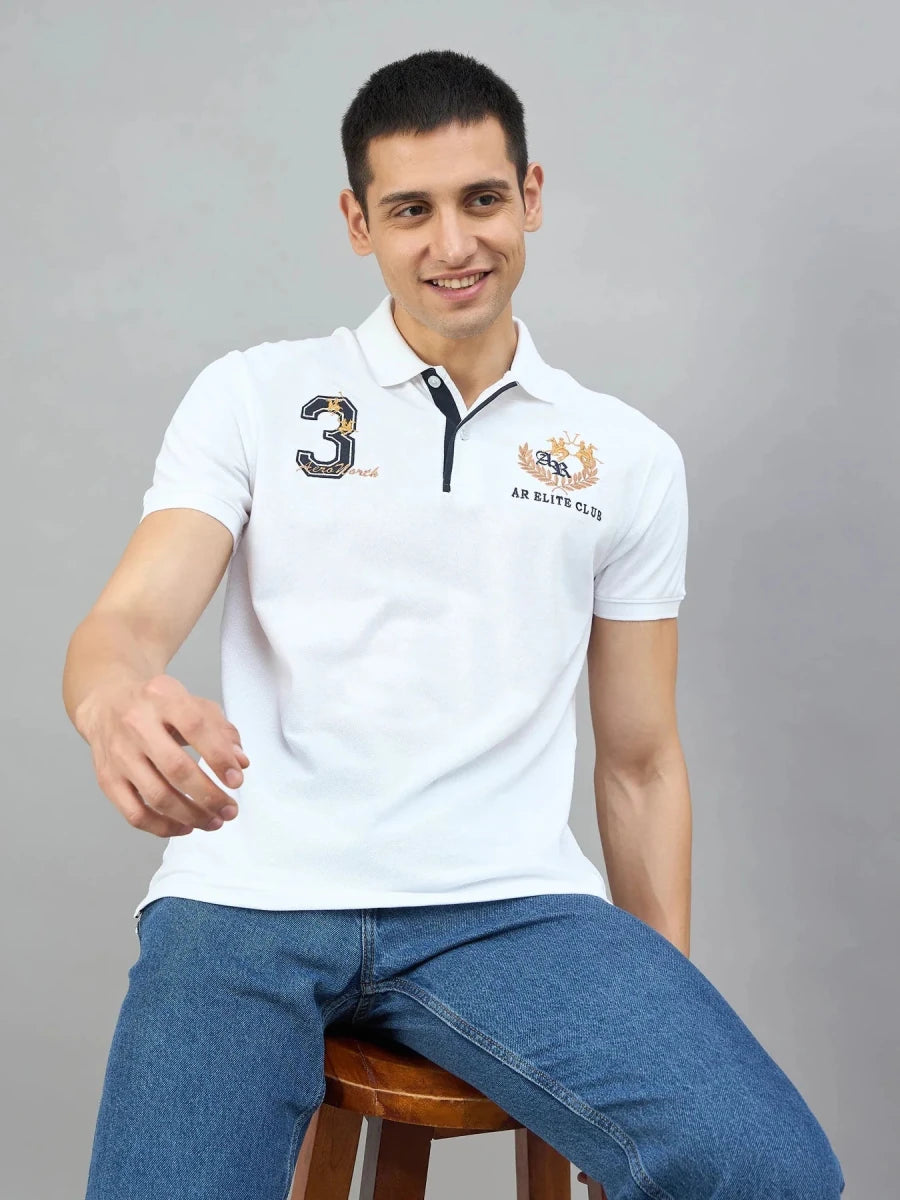 A man in a 98degreenorth Men's White AR Elite Club No.3 Polo T-Shirt sits on a stool, smiling against a gray background.