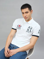 Man in a Men's White Embroidered Polo T-Shirt by 98degreenorth with blue jeans sits against a gray background.