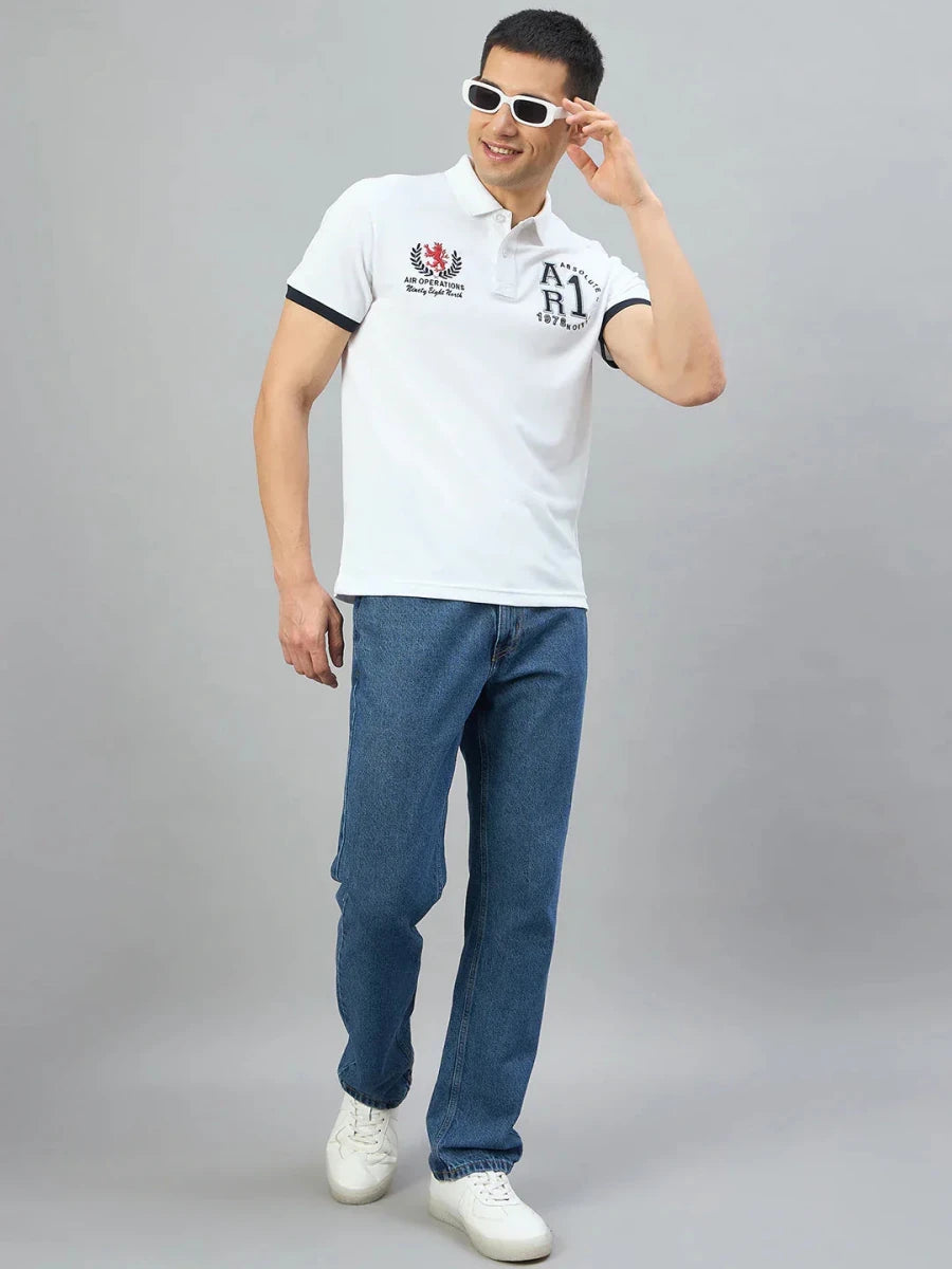 Man wearing 98degreenorth Men's White Embroidered Polo T-Shirt, blue jeans, and white sneakers on a gray background.