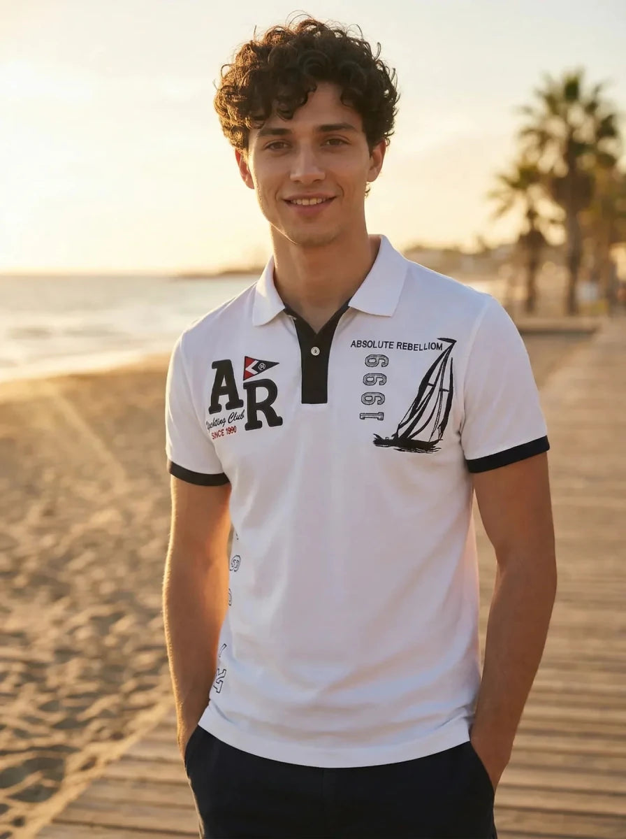 Smiling man wears the Men's White Yachting Club Polo T-Shirt by 98degreenorth on a beach boardwalk at sunset.