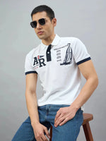 Man wearing sunglasses, blue jeans, and a 98degreenorth Men's White Yachting Club Polo T-Shirt, seated on a stool.