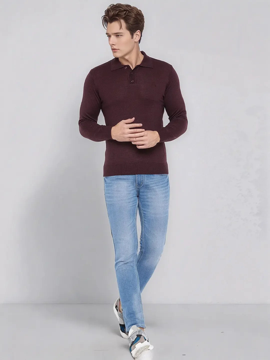A man wears a 98degreenorth Men’s Wine Merino Wool Polo Neck Sweater with light blue jeans against a gray background.