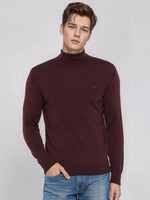 A man in a 98degreenorth men’s wine merino wool turtle neck sweater and blue jeans stands against a gray background.