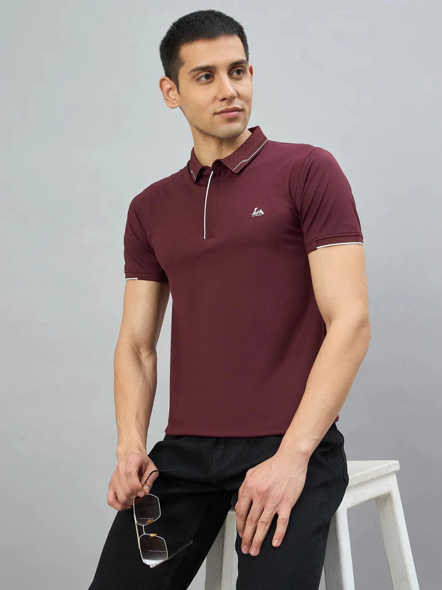 Man in a 98degreenorth Men's Wine Polo T-Shirt with White Tipping and black jeans, holding sunglasses, seated on a stool.