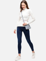 Woman in 98degreenorth Off White Ribbed Turtle Neck Sweater, smiling, walking in blue skinny jeans and sneakers.