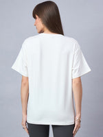 A woman wears the 98degreenorth Offwhite Typography Graphic Cotton Tee and black pants against a gray background.