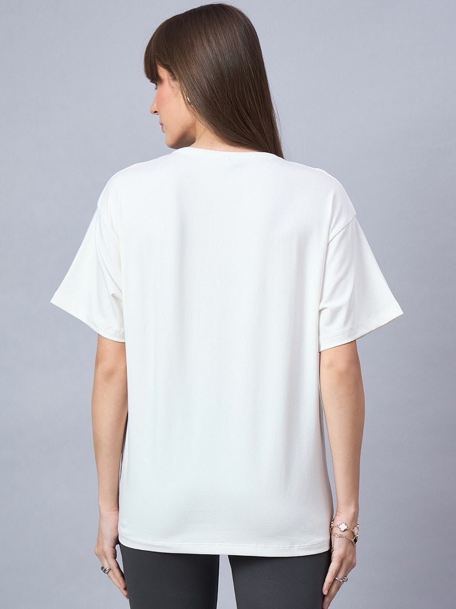 A woman wears the 98degreenorth Offwhite Typography Graphic Cotton Tee and black pants against a gray background.