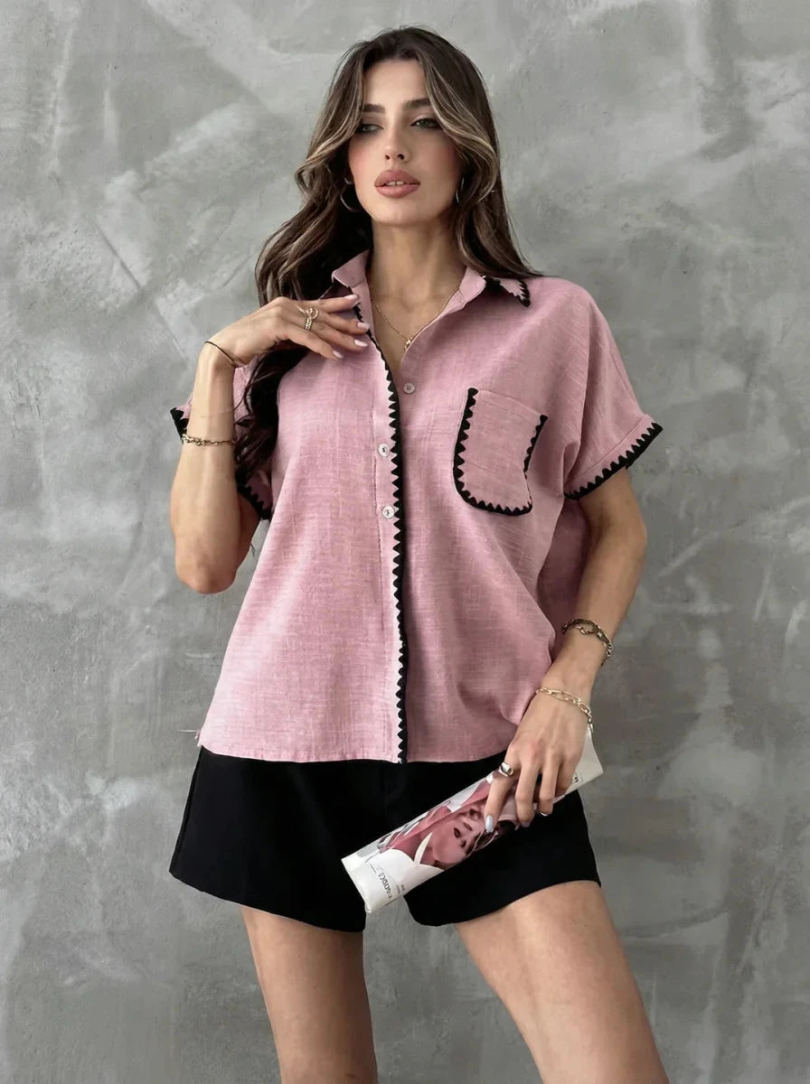 Woman in 98degreenorth's Pink Whipstitch Trim Cotton Shirt, holding a magazine, stands by a gray wall.