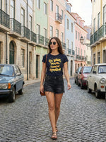 A woman in a 98degreenorth Regular Fit Cotton Typography T-shirt with a round neck walks past pastel buildings and parked cars.