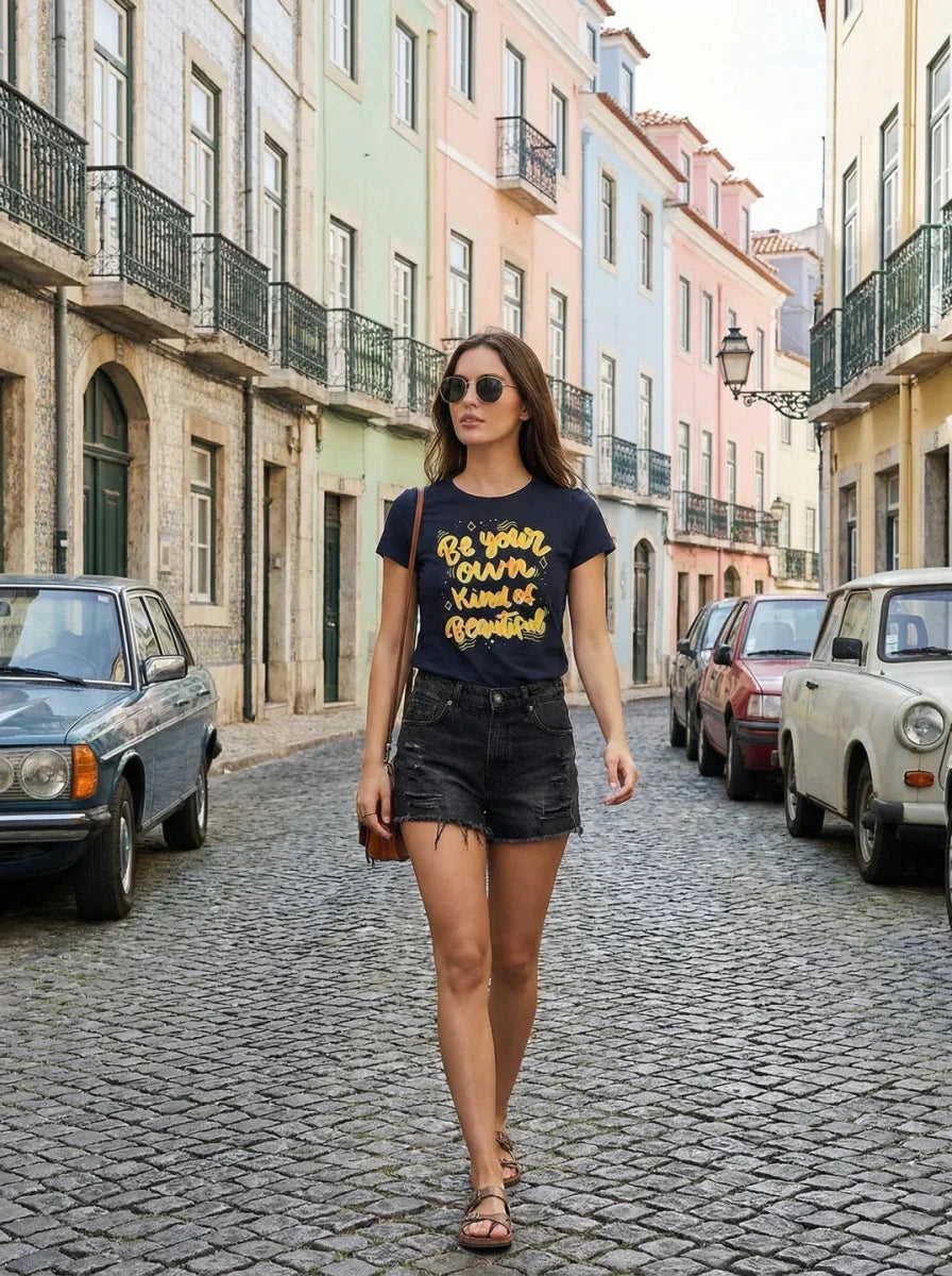 A woman in a 98degreenorth Regular Fit Cotton Typography T-shirt with a round neck walks past pastel buildings and parked cars.