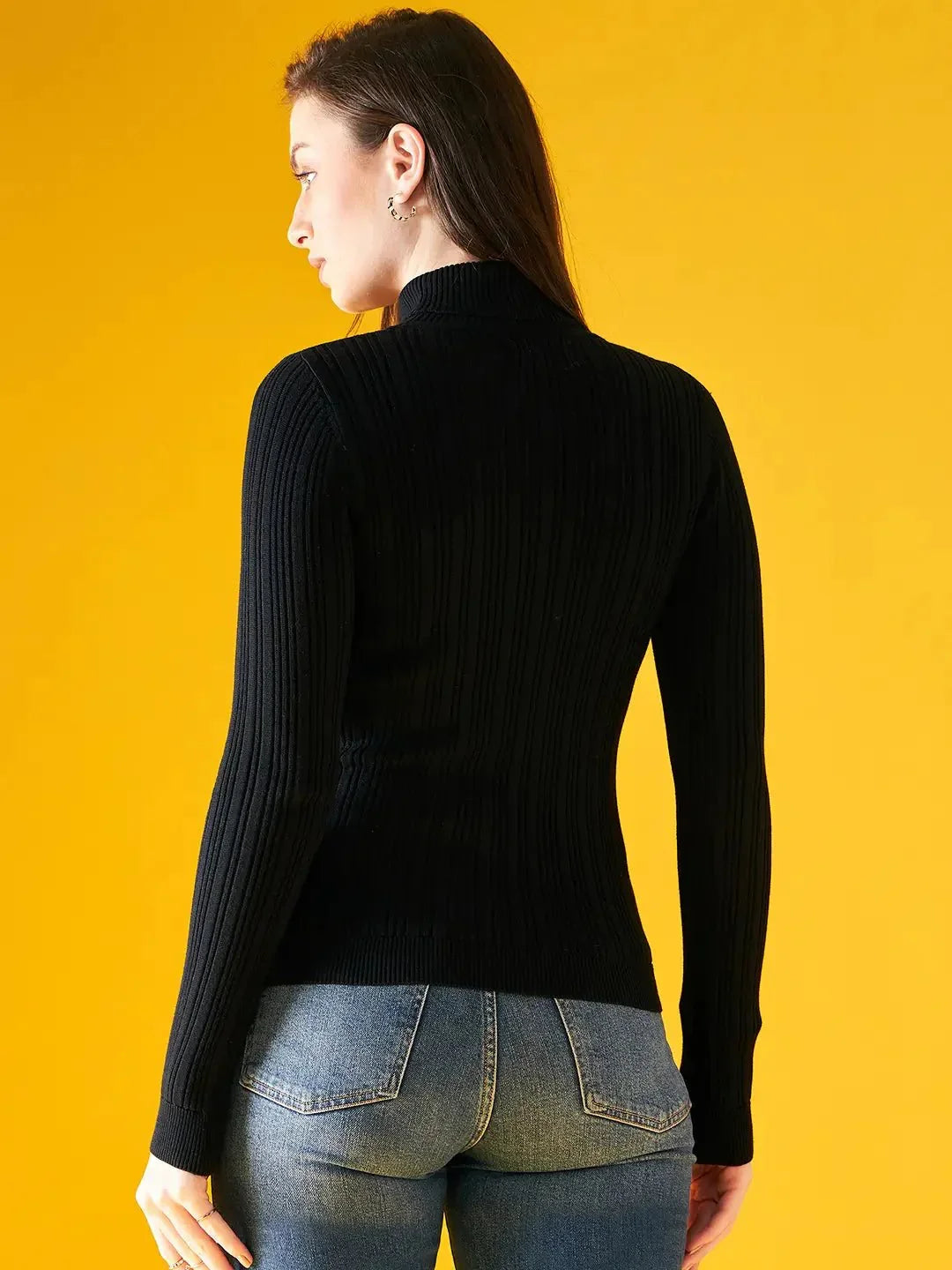 A woman with long brown hair, wearing the 98degreenorth Ribbed Black Turtle Neck Pullover and blue jeans, stands with her back to the camera against a solid yellow background.