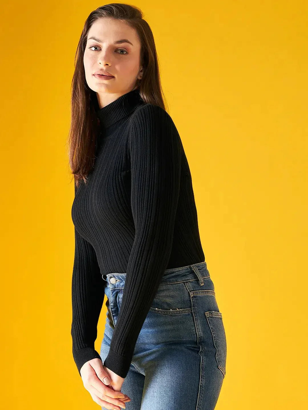 A woman with straight brown hair models the 98degreenorth Ribbed Black Turtle Neck Pullover with blue jeans against a bright yellow background.