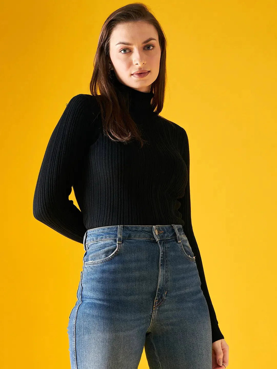 A person with long brown hair wears the 98degreenorth Ribbed Black Turtle Neck Pullover and blue high-waisted jeans, standing confidently against a solid yellow background and looking directly at the camera.