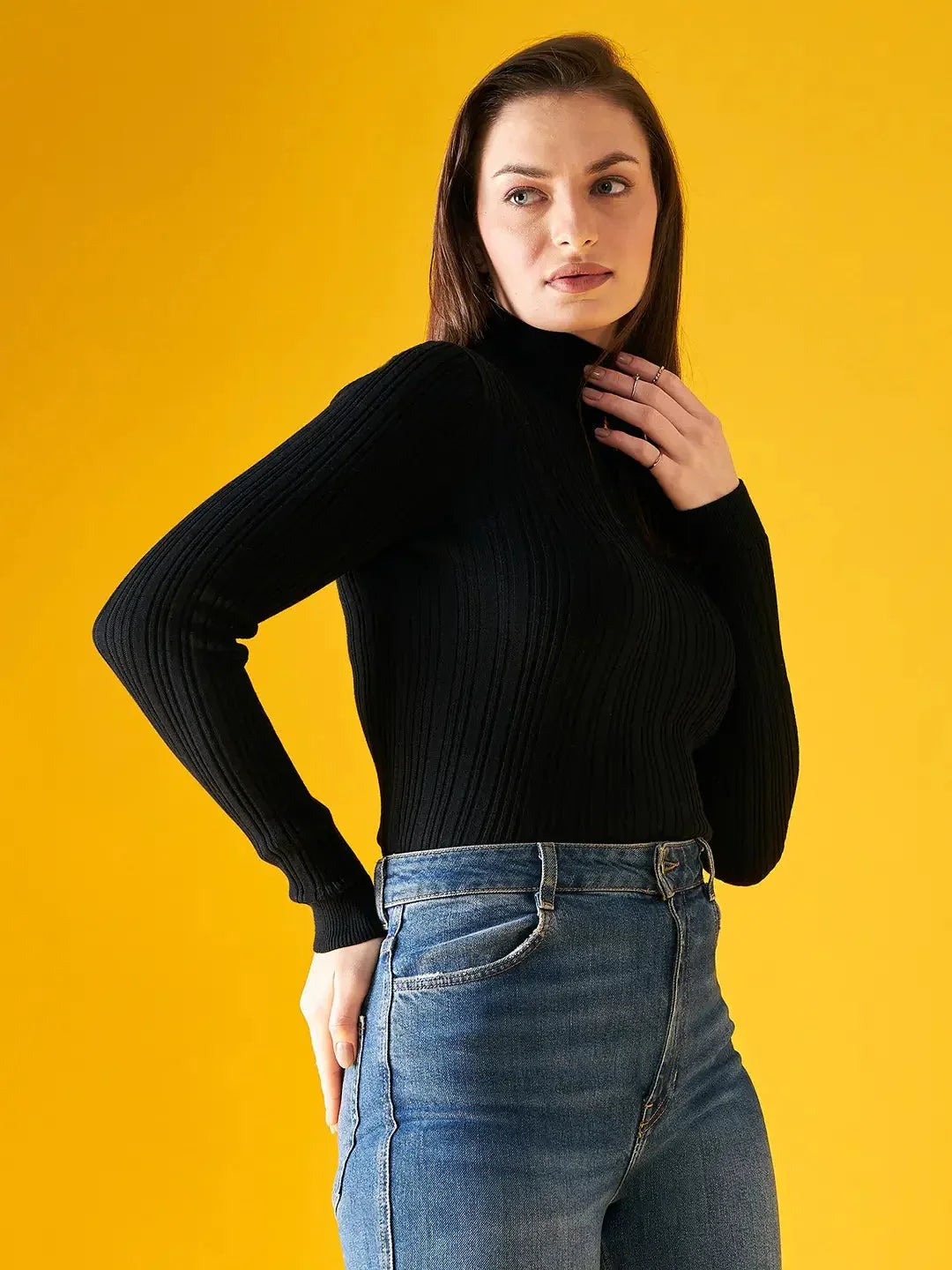 A woman wears the 98degreenorth Ribbed Black Turtle Neck Pullover with blue jeans, standing against a bright yellow background and gazing to the side, one hand near her neck and the other on her hip.
