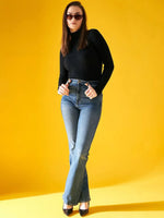 A woman with straight brown hair models the 98degreenorth Ribbed Black Turtle Neck Pullover, paired with sunglasses and blue jeans, standing confidently against a bright yellow background, hands on her hips.