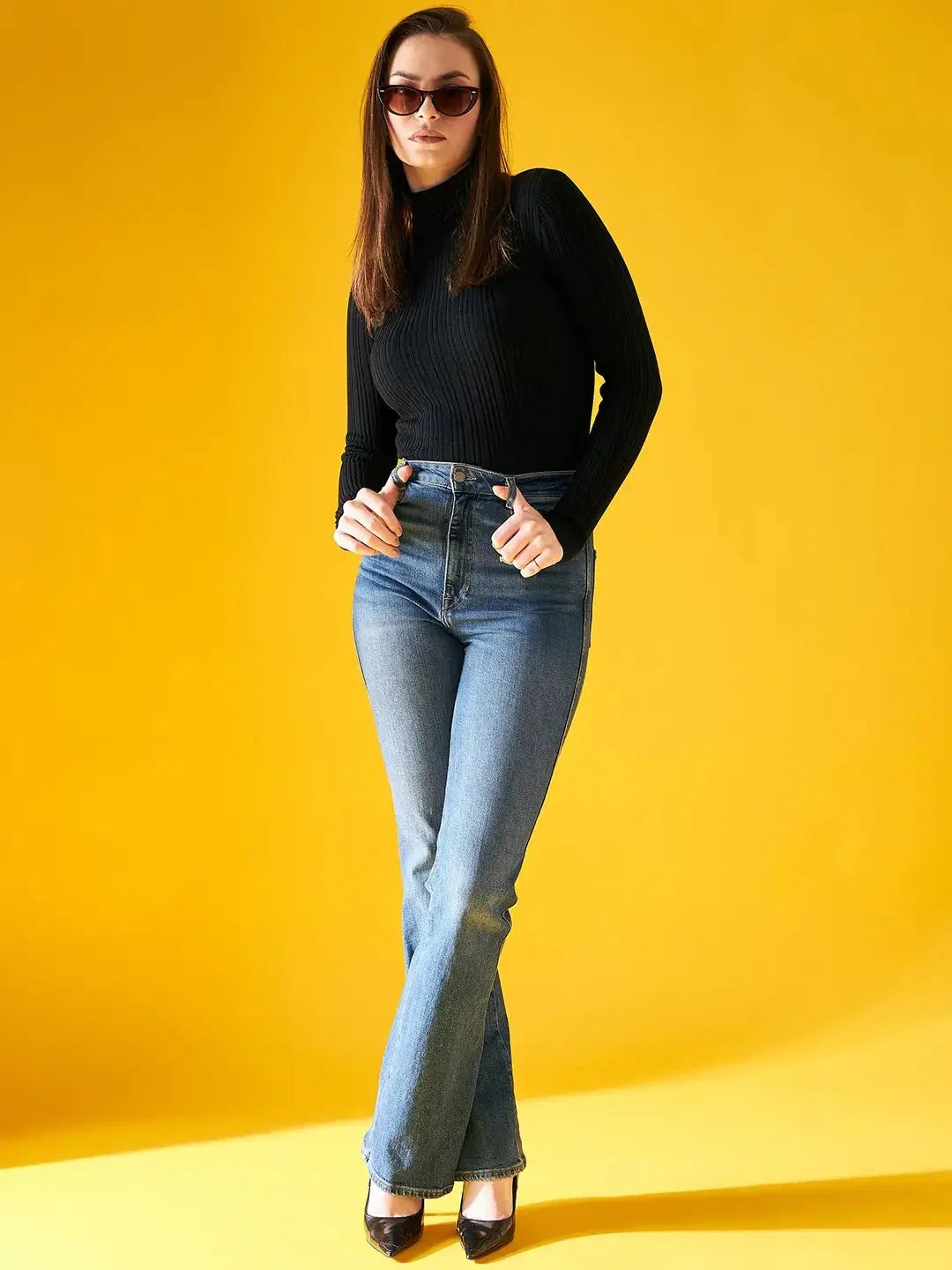 A woman with straight brown hair models the 98degreenorth Ribbed Black Turtle Neck Pullover, paired with sunglasses and blue jeans, standing confidently against a bright yellow background, hands on her hips.