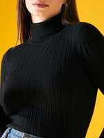 A woman wears the 98degreenorth Ribbed Black Turtle Neck Pullover with light blue jeans, standing against a bright yellow background. Her face is partially visible.