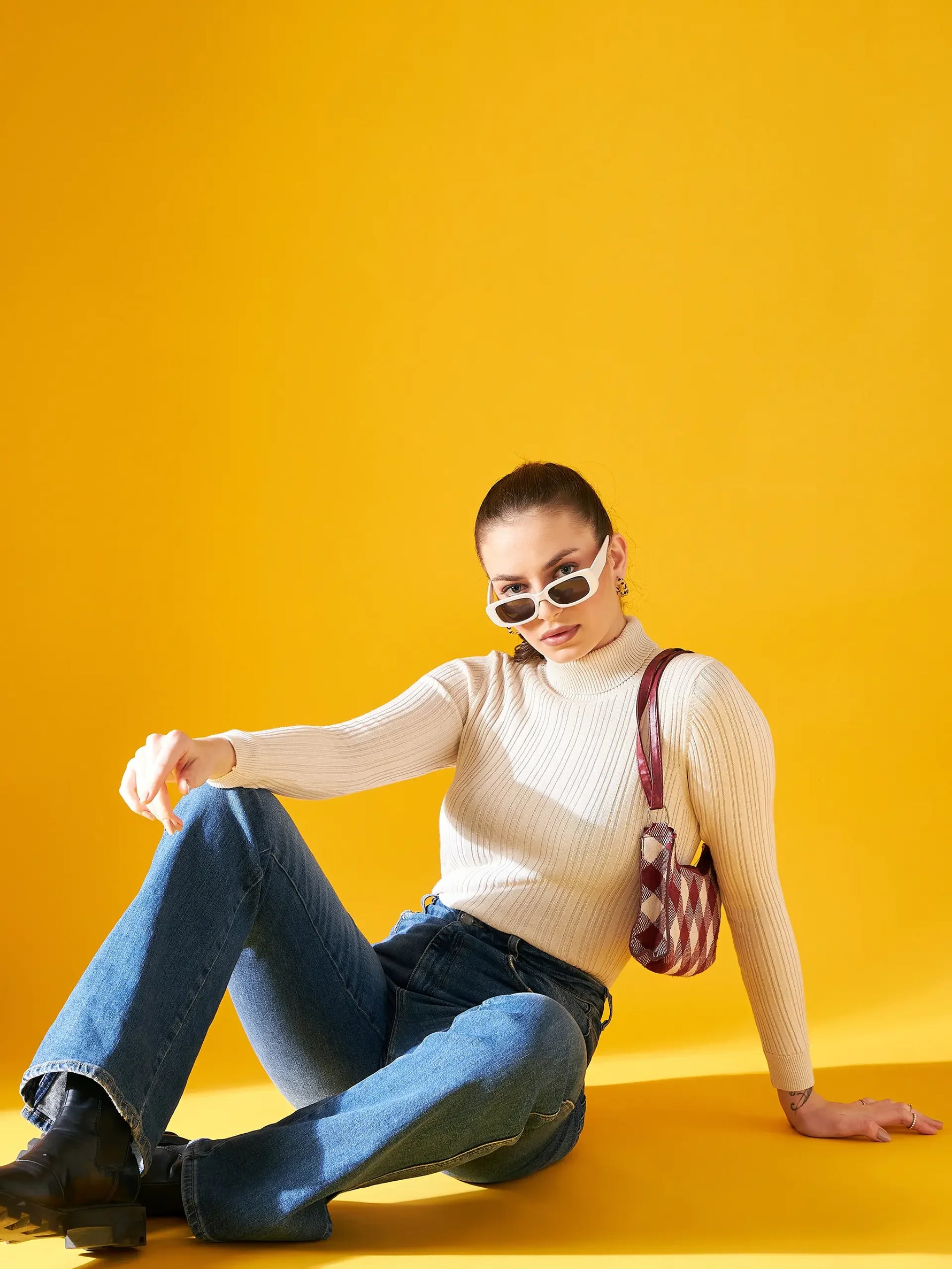 A woman sits on the floor against a bright yellow background, wearing white sunglasses, blue jeans, black boots, and a Ribbed Offwhite Turtle Neck Pullover by 98degreenorth with a small checkered shoulder bag. She looks confidently at the camera.