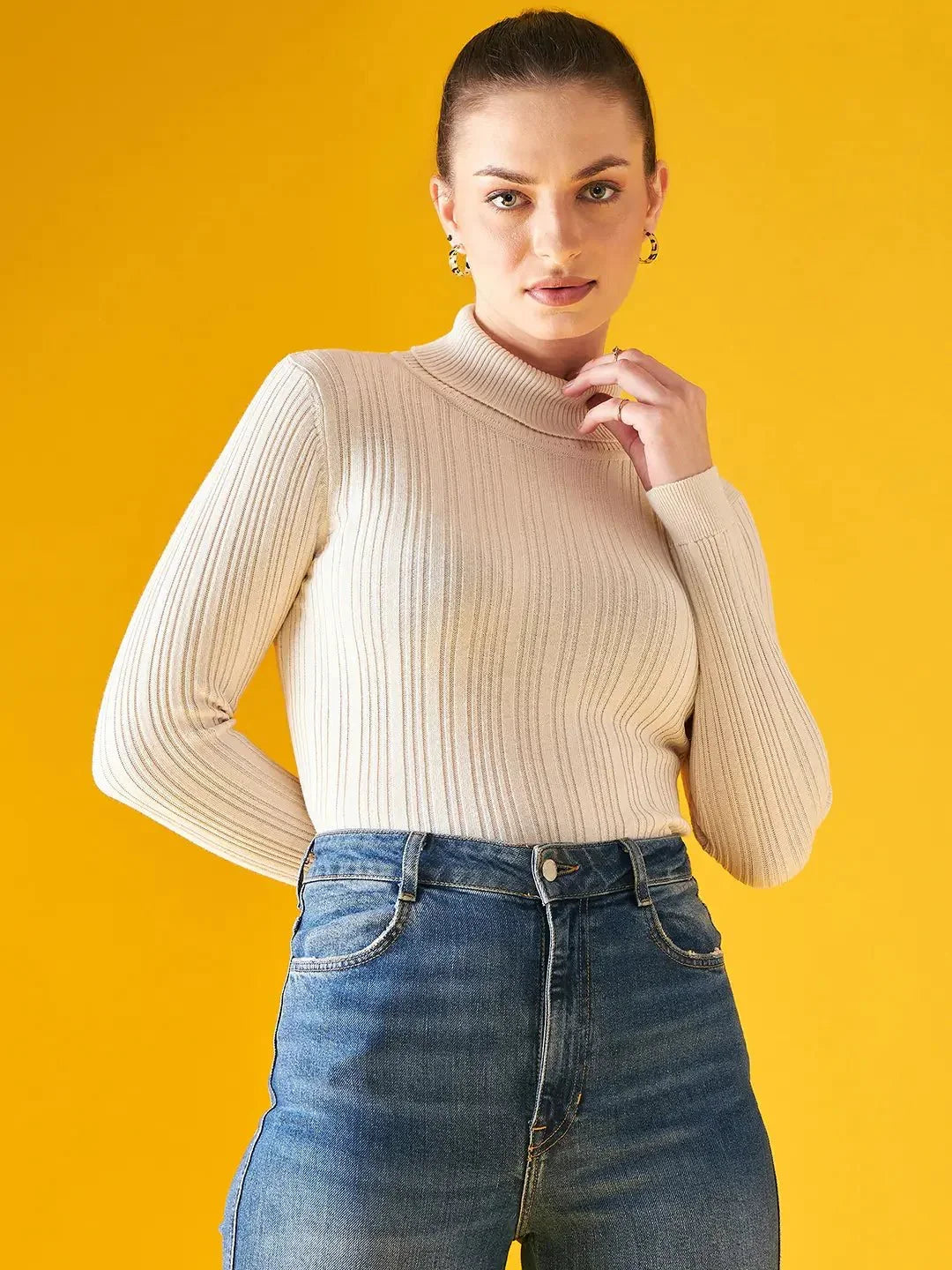 A woman with her hair tied back wears a 98degreenorth Ribbed Offwhite Turtle Neck Pullover and high-waisted blue jeans, standing against a solid yellow background. She looks at the camera with one hand near her chin.