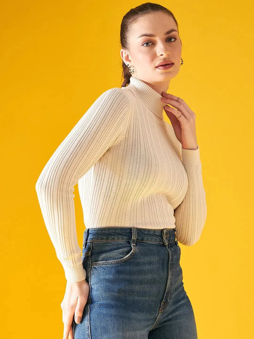 A woman in a 98degreenorth Ribbed Offwhite Turtle Neck Pullover and blue jeans stands against a bright yellow background, confidently looking at the camera with her hair pulled back.