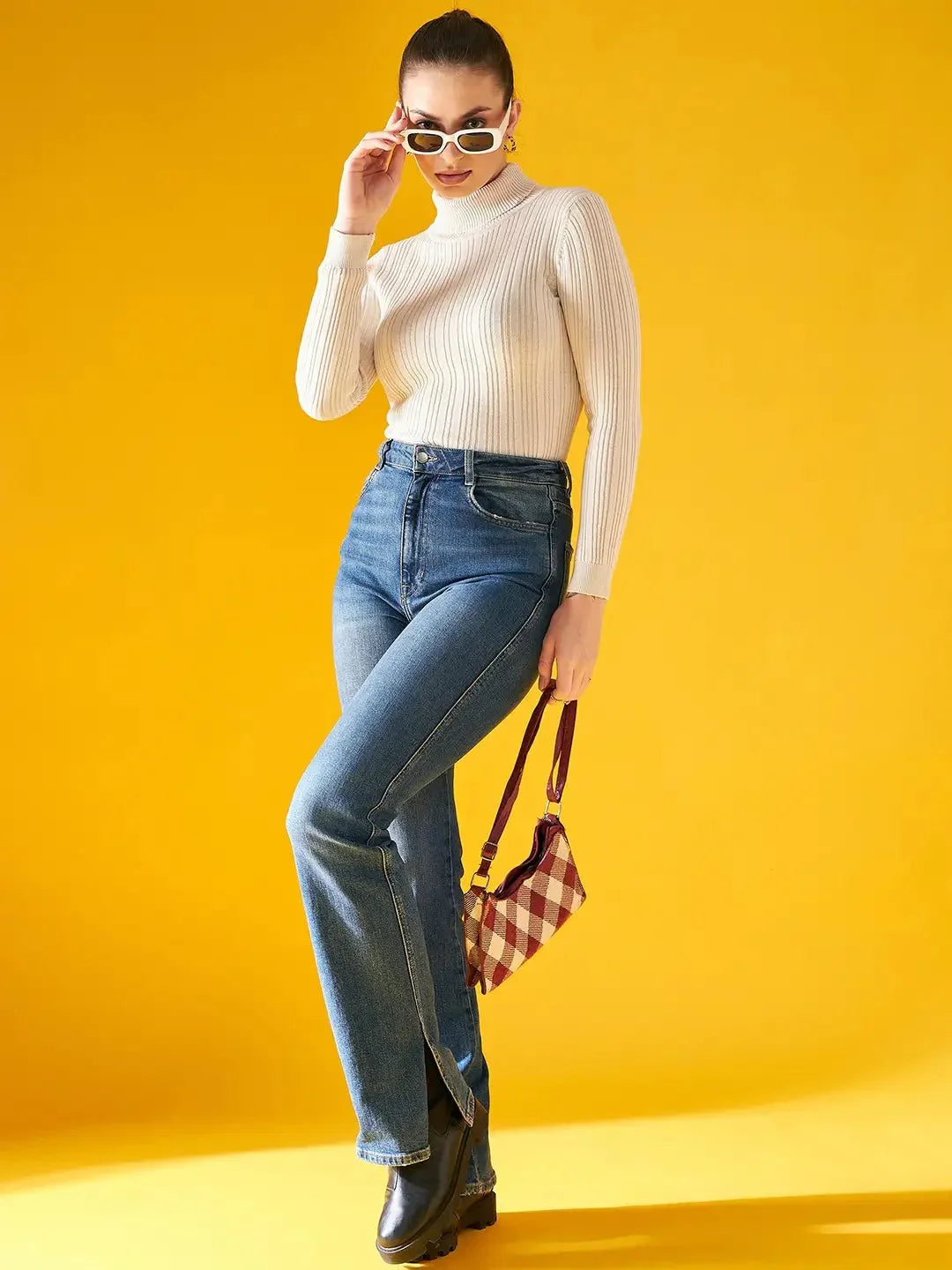 A woman poses confidently against a yellow background in a 98degreenorth Ribbed Offwhite Turtle Neck Pullover, high-waisted blue jeans, black boots, white sunglasses, and holding a small plaid handbag.