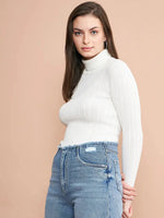 A woman with long brown hair wears the 98degreenorth Ribbed White Turtle Neck Pullover and high-waisted blue jeans made from premium cotton, standing against a beige background and smiling at the camera.