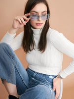 A woman in a 98degreenorth Ribbed White Turtle Neck Pullover and blue jeans sits against a beige background, looking to the side as she adjusts her large, square sunglasses.