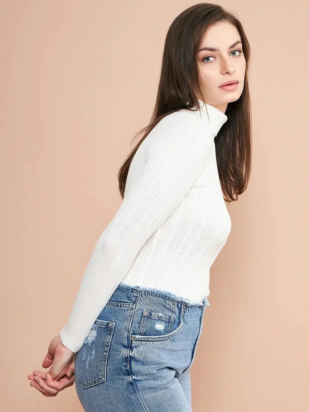 A woman with long brown hair models the 98degreenorth Ribbed White Turtle Neck Pullover with light blue distressed jeans, standing against a beige background with her hands clasped behind her back.