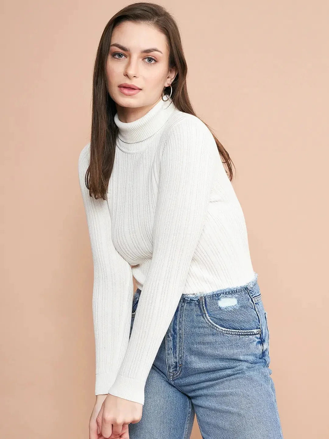 A woman with straight brown hair wears the 98degreenorth Ribbed White Turtle Neck Pullover and blue jeans, standing against a plain beige background with a relaxed expression.