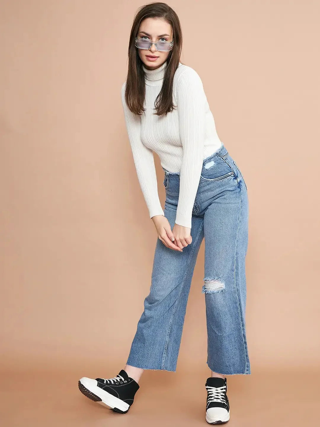 A woman in a 98degreenorth Ribbed White Turtle Neck Pullover, blue ripped high-waisted jeans, black-and-white sneakers, and blue-tinted sunglasses poses against a beige background.