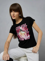 A woman in a black 98degreenorth round-neck typography t-shirt and white pants sits on a stool against a plain gray background.