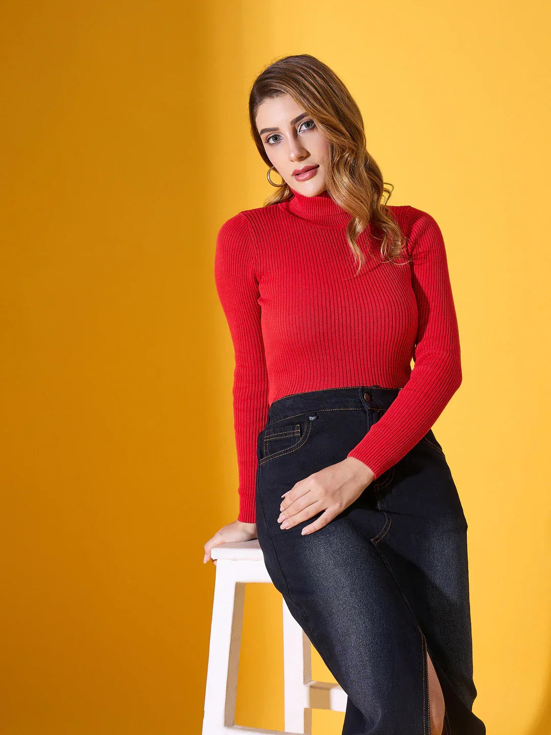 Woman wears 98degreenorth Scarlet Red Ribbed Turtleneck Sweater and dark jeans, sitting on a stool against yellow.