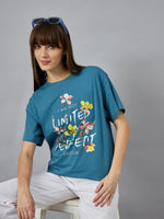 Woman wears 98degreenorths denim Stylish Typography Printed T-shirt and white pants, seated against a gray background.