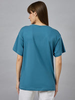 A woman with straight brown hair wears a 98degreenorth drop shoulder typography t-shirt and white pants, facing away.