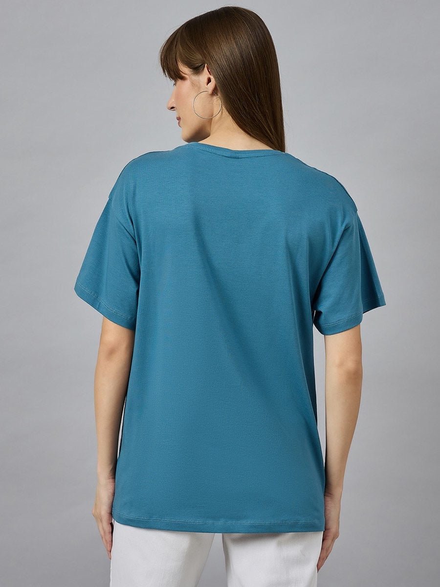 A woman with straight brown hair wears a 98degreenorth drop shoulder typography t-shirt and white pants, facing away.