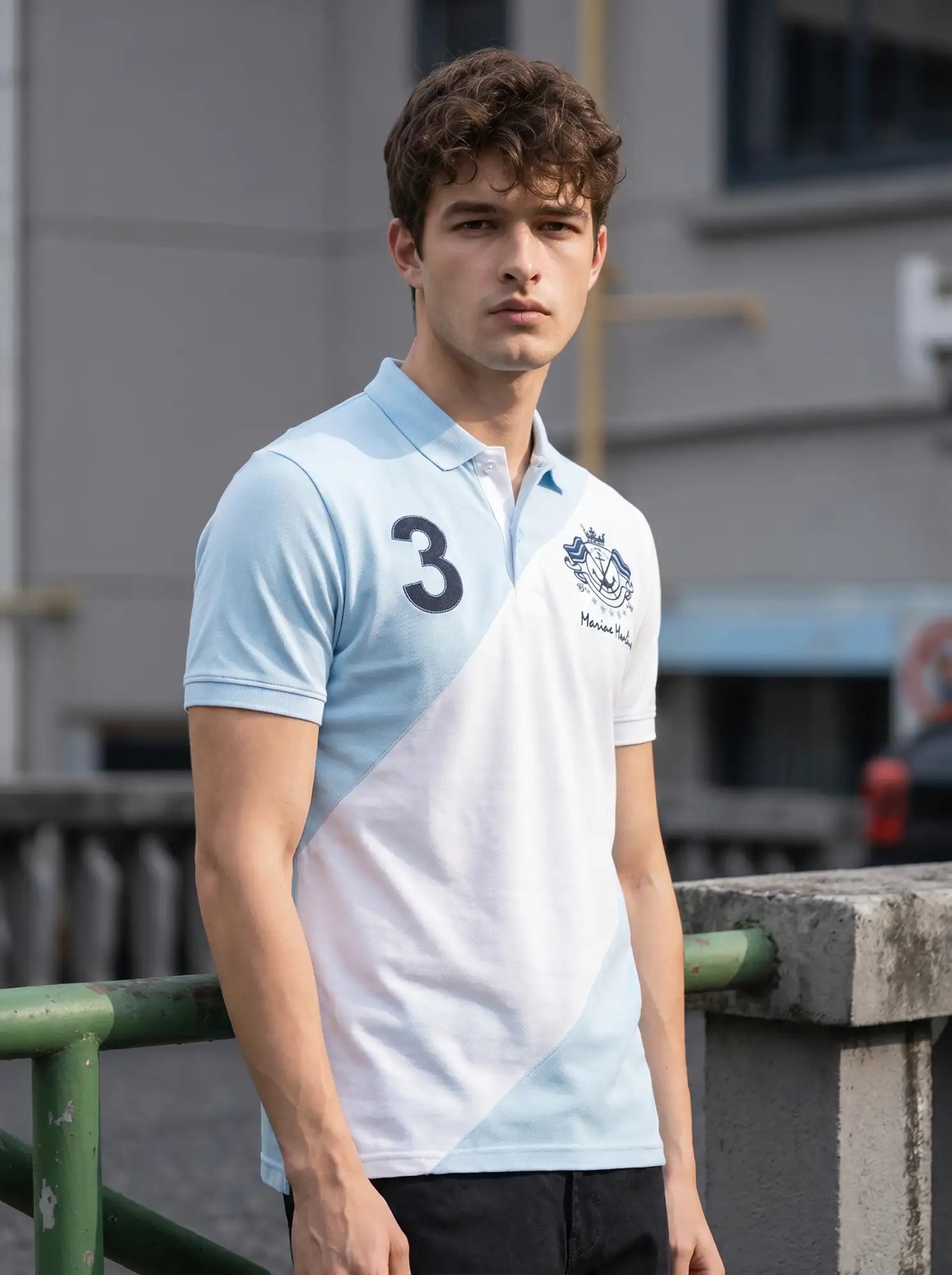A man in a 98degreenorth sky blue & white colour block polo stands by a green railing, looking at the camera.