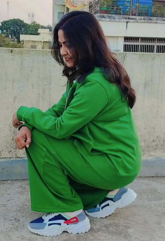 Woman in a green outfit squatting on a rooftop, looking down and smiling, with long dark hair and colorful sneakers.
