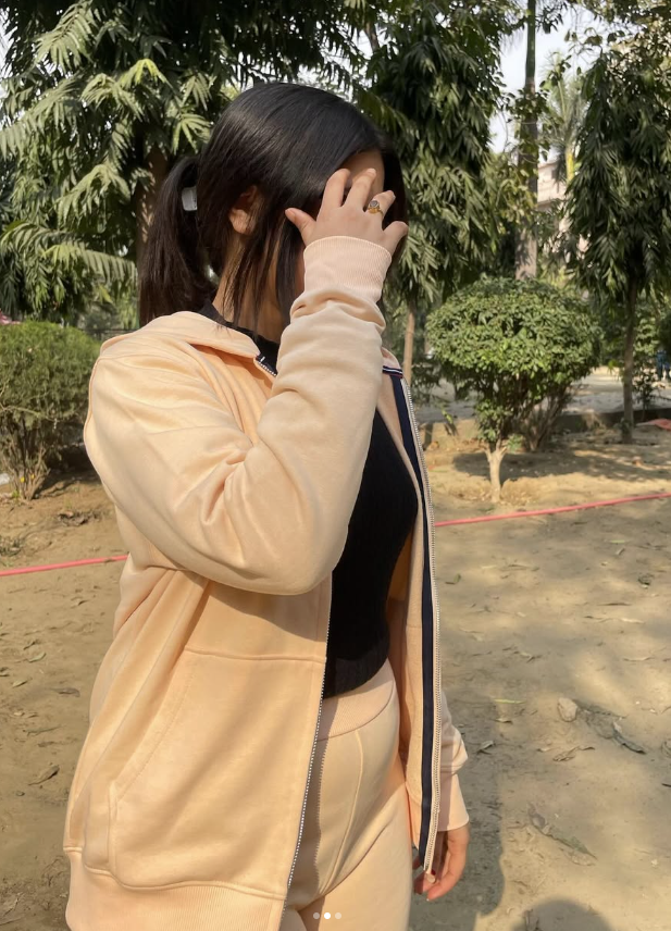 A person in a peach tracksuit covers their face with one hand while standing outdoors in a sunny park.