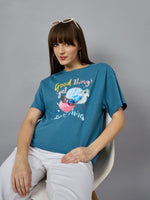 A woman in a 98degreenorth Trendy Typography Graphic T-shirt sits on a white chair, styled with white pants and sunglasses.