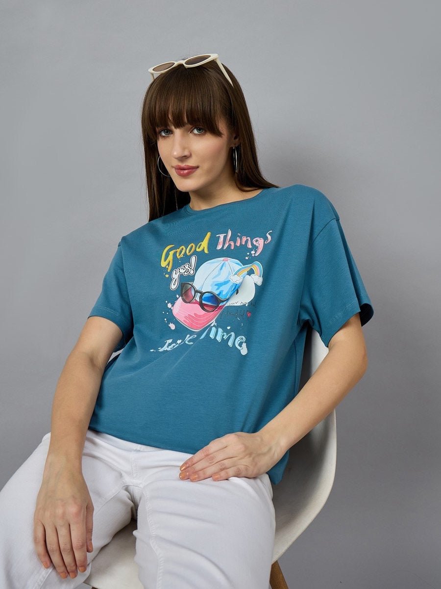 A woman in a 98degreenorth Trendy Typography Graphic T-shirt sits on a white chair, styled with white pants and sunglasses.