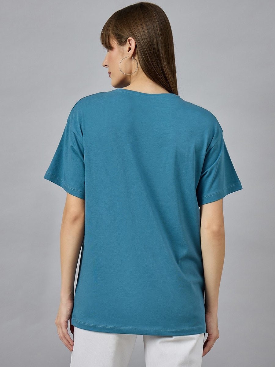 Woman with straight brown hair in a 98degreenorth Trendy Typography Graphic T-shirt, white pants, and hoops, facing away.