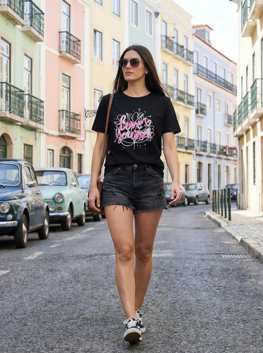 Woman in a 98degreenorth Typography Graphic Cotton T-shirt, sunglasses, and shorts walks a city street with vintage cars.