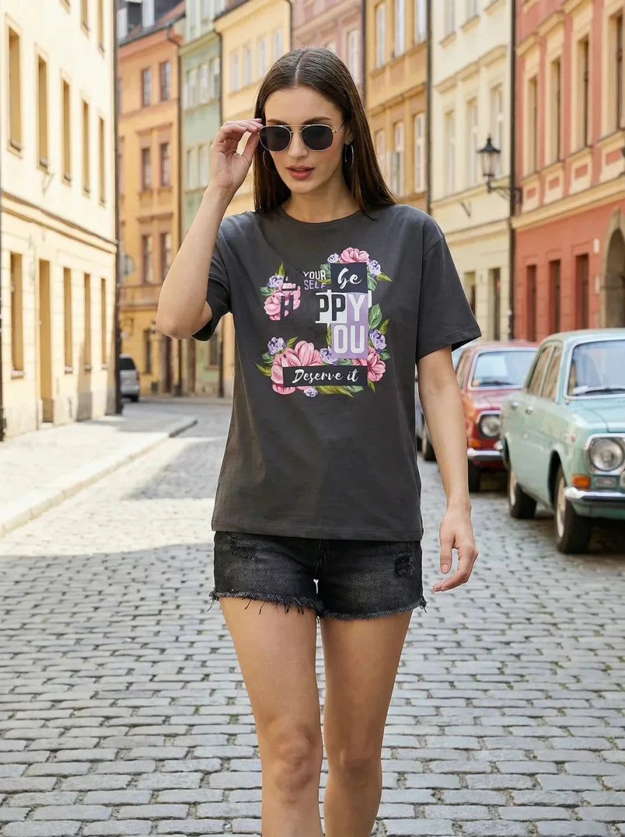 Woman in 98degreenorth Typography Graphic Cotton Tee strolls a cobblestone street lined with colorful buildings.