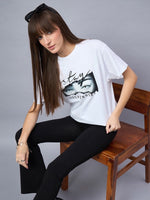 Woman with long brown hair wears a 98degreenorth Typography Print Drop Shoulder T-shirt, seated on a wooden chair.