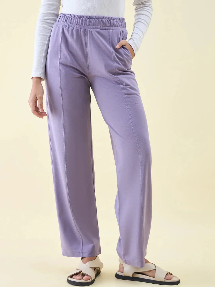 Person in 98degreenorth Violet Wide Leg Pintuck Trackpants, white long-sleeve top & beige sandals, hands in pockets.