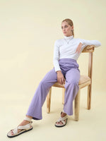 A woman in a white turtleneck and 98degreenorth Violet Wide Leg Pintuck Trackpants sits on a wooden chair.